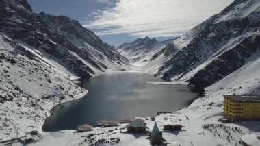 An aerial shot of a lake surrounded by snow coverred mountains on a cloudy day