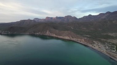 An aerial view of the wavy sea and the rocky mountains gleaming under the cloudy blue sky in 4K