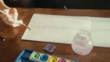 A closeup shot of a female hand drawing on a white paper with a painting brush near palette of colors on a wooden table
