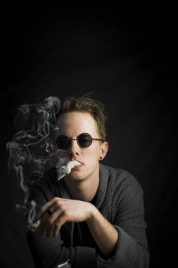 The vertical shot of a smoking young guy wearing round black sunglasses isolated on the black background