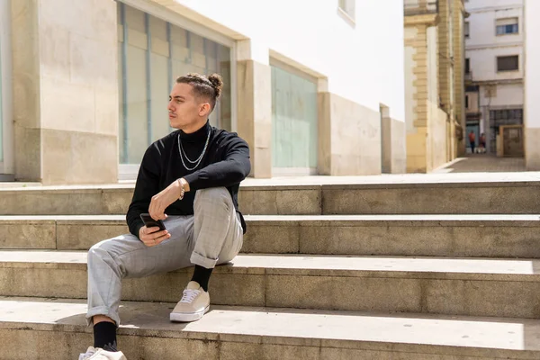 Genç bir melez adamın portresi. Şehirde merdivenlerde oturan akıllı bir telefonu düşünceli bir şekilde tutuyor. Sosyal medya, commu, mobil uygulamalar, yaşam tarzı kavramı