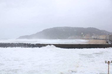 Ormanla kaplı tepe manzarası ve sisli bir havada kumsaldaki binalar.