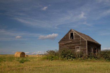 Bir tarladaki ahşap bir çiftlik evinin panoramik görüntüsü. Bulutlu bir gökyüzünün altında sarı, kuru otlar arasında. Manitoba, Kanada