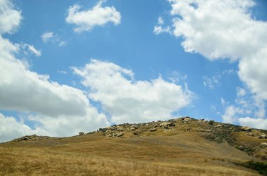 Baharda beyaz bulutlarla dolu mavi gökyüzünün altına dağılmış yeşil bir tepe manzarası.