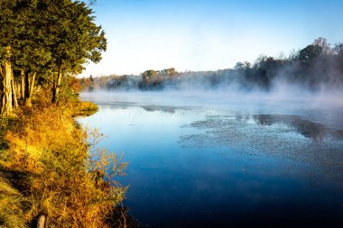 Güneşin battığı sisli bir günde, sonbahar renkli ağaçlarla çevrili sakin bir göl manzarası.