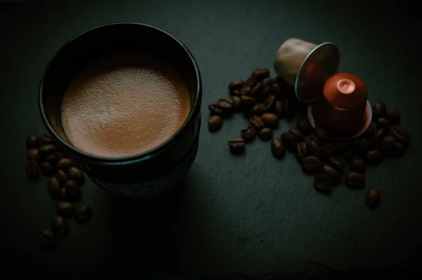 A cup of coffee with capsules and beans on a black table