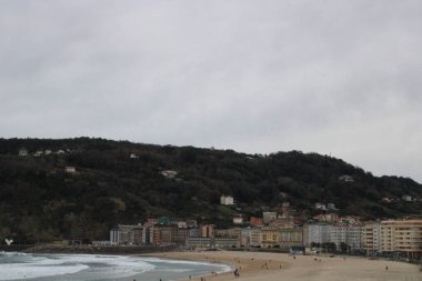 Ormanla kaplı tepenin manzarası ve bulutlu gökyüzünün altındaki kumlu sahildeki binalar.