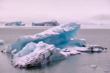 Fjallsarlon buzulu gölünde yüzen büyük mavi ve siyah buzdağı ve arka planda diğer büyük buzdağları.