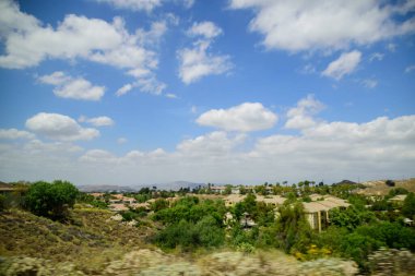 Parlak mavi gökyüzünün altında yeşil çalılarla ve alçak binalarla kaplı bir alan manzarası