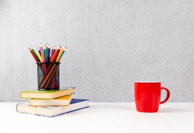 Renkli kalemlerle dolu bir yığın kitap ve beyaz bir masanın üzerinde bir fincan kahve.