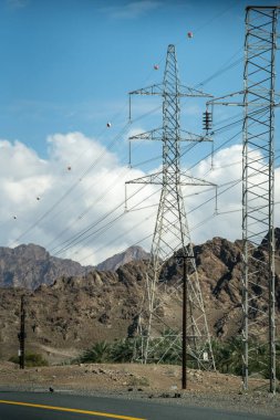 Yol kenarındaki elektrik kablolarının dikey görüntüsü. Parlak güneş ışığında kayalık dağlarla mavi bulutlu gökyüzü karşı karşıya.