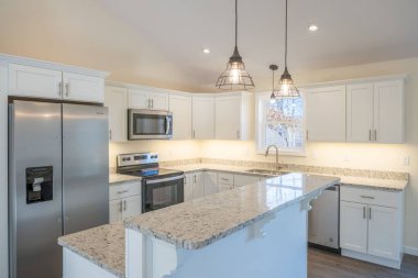 Tavanda asılı beyaz dolaplar, buzdolabı, fırın ve lambalar olan rahat, modern bir mutfak.