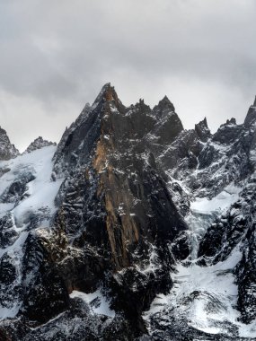 Gri bulutlu gökyüzünün altında karla kaplı bir dağın yüksek manzarası.