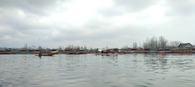 Göldeki teknelerin panoramik bir görüntüsü, bulutlu bir gökyüzüne bakan ağaçların yansıması.