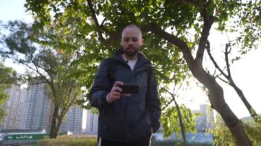 Parkta beyaz bir erkeğin yakın çekimi. Bir şey söylüyor, telefonuna bakıyor.