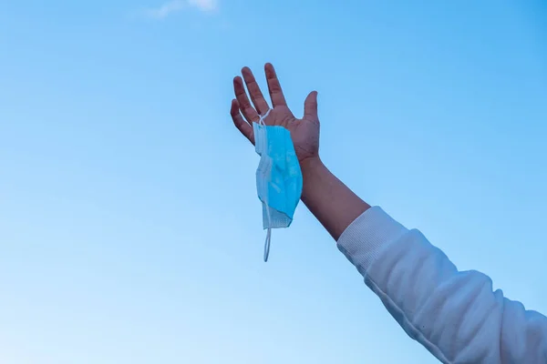 A hand raising a mask with the sky in the background, end of the obligatory use of masks.