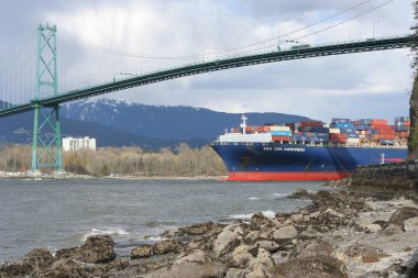 Köprünün arka planında denizde yüzen bir konteyner gemisinin yatay görüntüsü.
