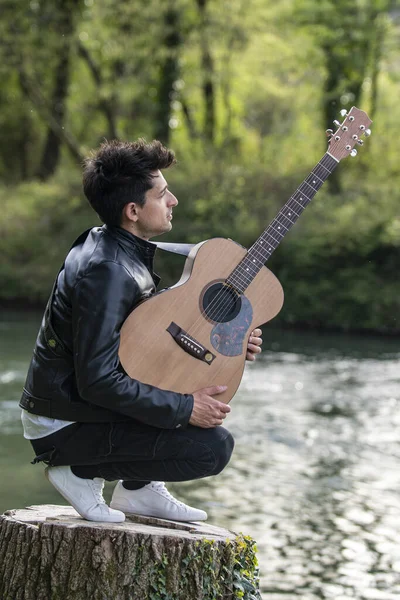 A vertical shot of a caucasian man with a guitar kneeing on a wooden stump by the lake