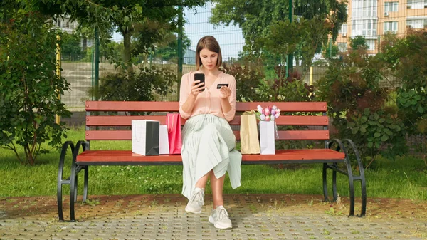 白人年轻女子坐在公园的长椅上 提着购物袋 拿着信用卡和手机 — 图库照片