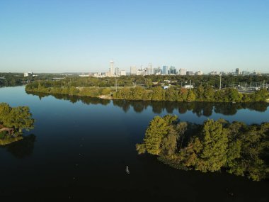 Bir nehrin kuş bakışı manzarası kıyılarındaki yeşil ağaçları mavi gökyüzünün altındaki şehir merkezine yansıtıyor.