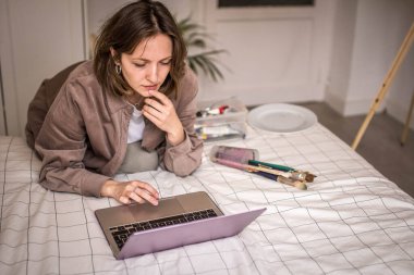 Çırak ressamın resmi ilham alıyor Laptop 'la fikir alıp fırçayla fırçaları fırçayla boyarken odasında Easel' le