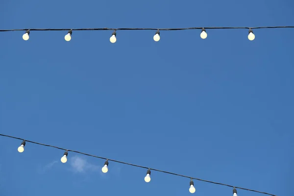 A low angle shot of a string of lit decorative light bulbs in the street against a blue sky