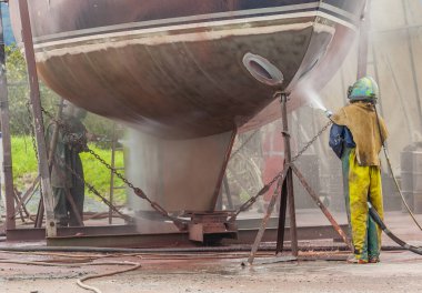 İşçi, yüksek basınçlı kum püskürtme sistemiyle bir yelkenli gemisinin gövdesini kumla kaplıyor.