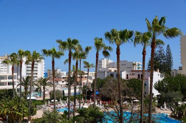 Resim, Cala Millor Mallorca 'da bahsi geçen hipotellerin havuz alanındaki panoramik manzarayı ve arka planda büyük palmiye ağaçları ve binaları gösteriyor.