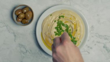 Bir insanın elinin en üst görüntüsü nar tohumlarını beyaz bir mutfak mermerinin üzerindeki yeşil zeytin tabağının yanındaki humus tabağına koyar.