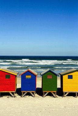 Güneşli bir günde berrak bir gökyüzünün altında sakin bir deniz manzarası olan canlı, renkli küçük plaj kulübelerinin dikey görüntüsü.
