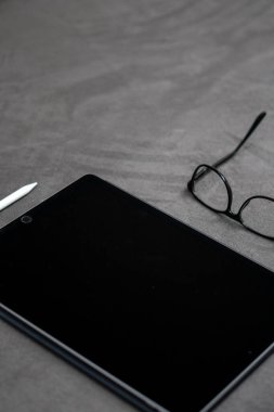 Gri kumaşın üzerinde beyaz kalem ve gözlüklü siyah ince bir tabletin dikey yakın çekimi.