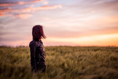 Deri ceketli genç kadın yeşil tarlada duruyor güzel günbatımının tadını çıkarıyor..