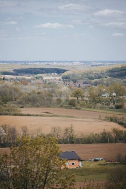 Kırsal kesimde bulutlu bir gökyüzünün altında yeşilliklerle çevrili bir kasabanın dikey görüntüsü.