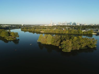 Bir nehrin kuş bakışı manzarası kıyılarındaki yeşil ağaçları mavi gökyüzünün altındaki şehir merkezine yansıtıyor.