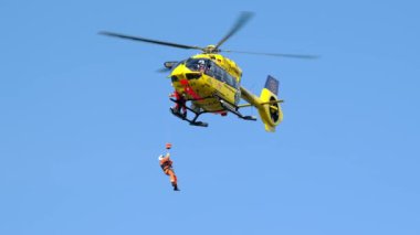 Mavi gökyüzünün önünde uçan bir ADAC kurtarma helikopterinden bir kişi çekiliyor. Alman hava ambulansı, arama kurtarma sistemi.