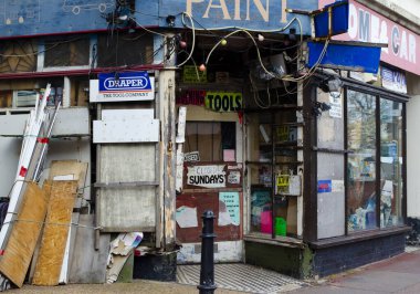 Birden fazla parlak ve yırtık tabelaları olan eski moda bir hırdavatçı. Dış tarafı solmuş ve pul pul dökülmüş boya ve eski kablolar ile aşınmış..