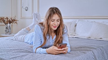 Kafkasyalı genç kadın pijama giyiyor ve yatakta yatarken telefonunu kontrol ediyor.