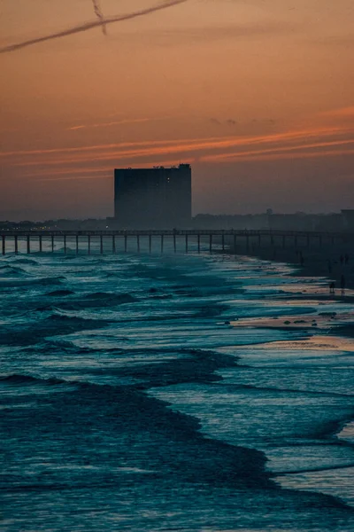 A vertical shot of waves washing the shore with a building silhouette in the background at dusk