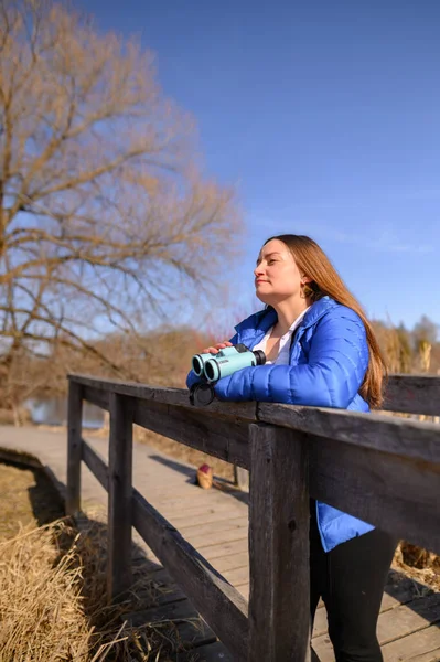 A woman in a blue down jacket looks into the distance on a bridge holding binoculars in a park on a sunny spring day.