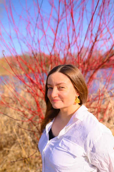 Portrait of a woman with a slight smile looking at the camera standing in front of a red dogwood bush with a river behind it during the day
