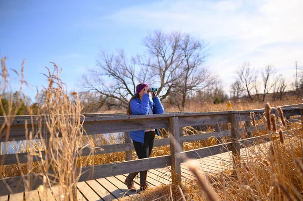 A woman in a blue down jacket looks into the distance through blue binoculars on a bridge in a park on a sunny spring day.