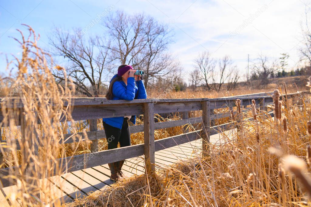 A woman in a blue down jacket looks into the distance through blue binoculars on a bridge in a park on a sunny spring day.