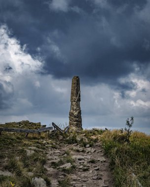 Kasvetli bir günde, koyu mavi bulutların altında, çimenli bir tepedeki eski, uzun bir kayanın dikey görüntüsü.