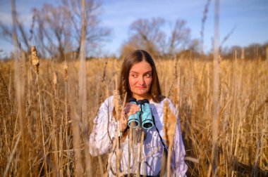 Kafası karışık bir ifadeyle gün boyunca kedi kuyruğu tarlasında duran mavi dürbünle yan tarafa bakan bir kadın..