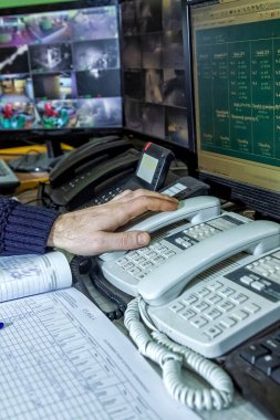 Yanında başka telefonlar olan bir adamın elinin dikey görüntüsü, arka plandaki bilgisayarlar ve diğer iş yeri ekipmanları.