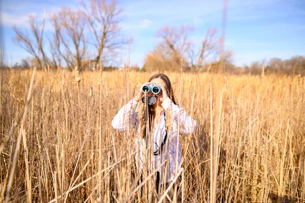Profile of a woman looking to the side through blue binoculars standing in a field of cat tails in a marsh during the day.