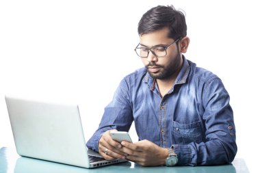 Beyaz arka planda dizüstü bilgisayarın önünde cep telefonuyla yazı yazan dikkati dağılmış genç bir adam.