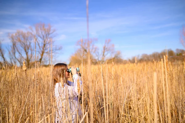 Profile of a woman looking to the side through blue binoculars standing in a field of cat tails in a marsh during the day.