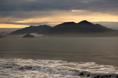 Gün batımında bulutlu bir gökyüzünün altında siluet dağları olan bir deniz pelerini.