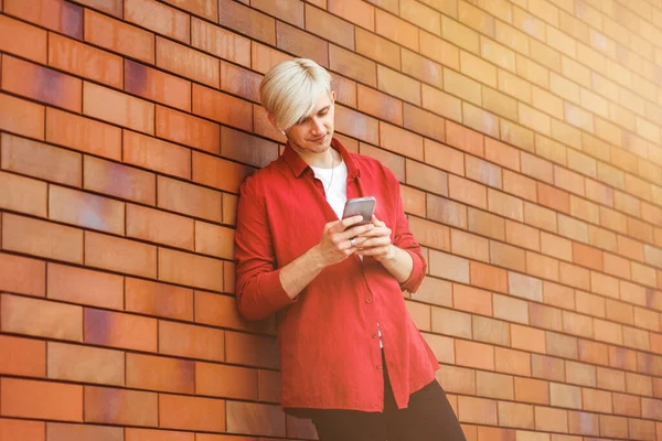 A young blonde Caucasian guy looking at his smartphone near a brick wall